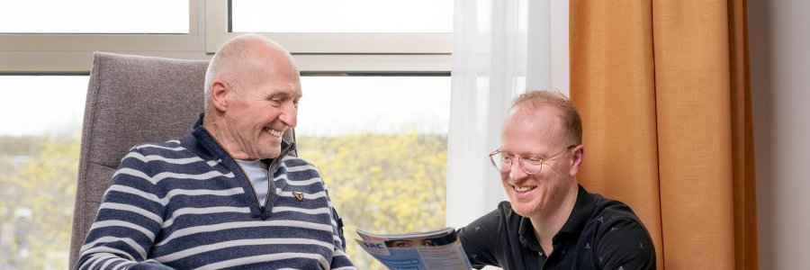 Een meneer zit op een stoel terwijl een andere persoon naar een magazine kijkt - Uitgesneden