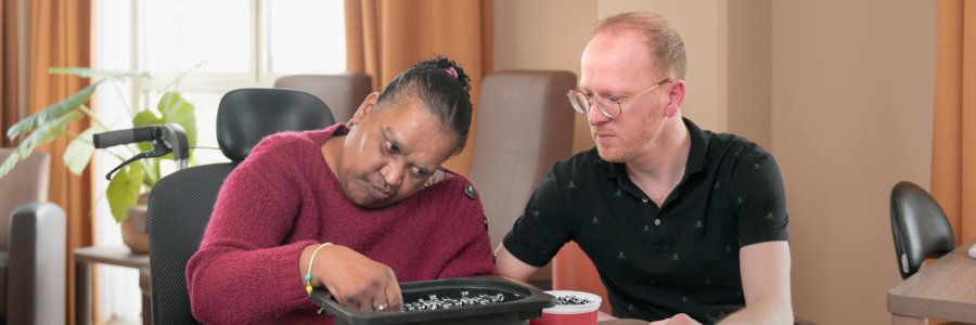 Een mevrouw zit schroeven en bouten te sorteren uit bakje en een meneer naast haar begeleid haar daar mee 