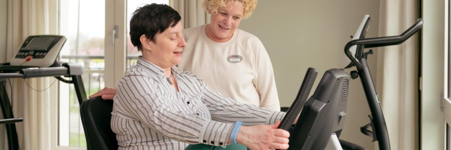 Een vrouw zit op een fitness fiets terwijl iemand anders haar begeleid 