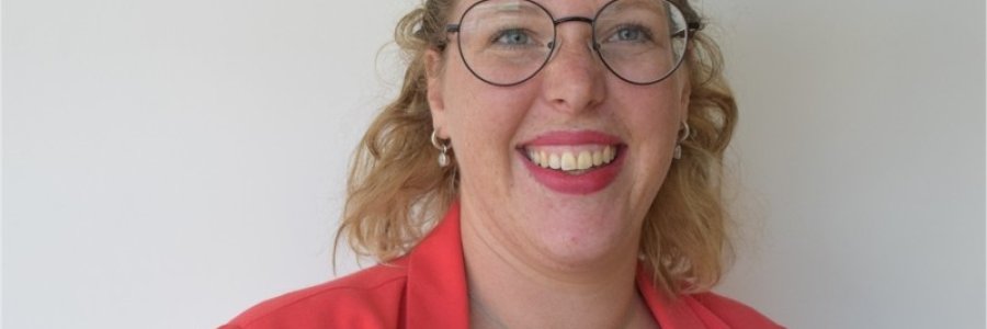 Een portretfoto van Marieke, lachende witte blonde vrouw met krulletjes, bril en rood jasje