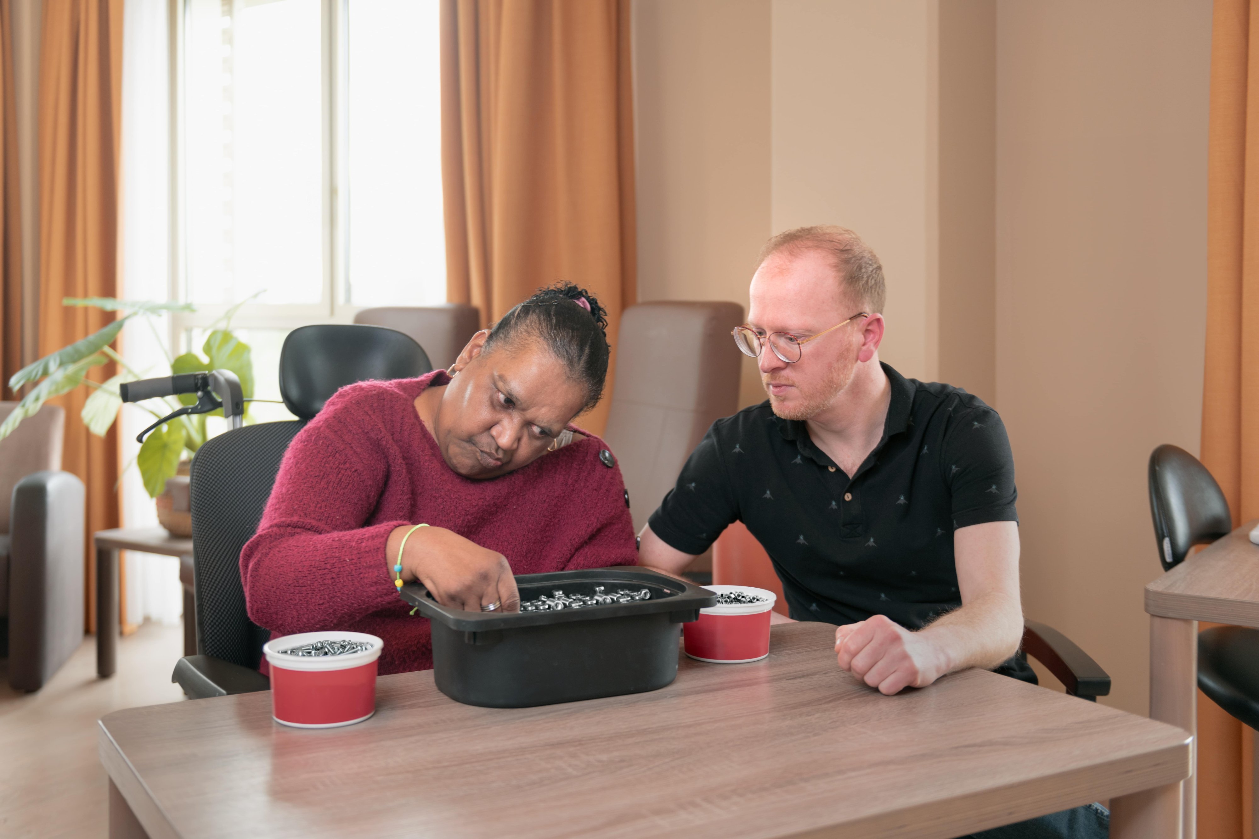 Een mevrouw zit schroeven en bouten te sorteren uit bakje en een meneer naast haar begeleid haar daar mee 